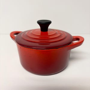 May include: A small, round, red enameled cast iron Dutch oven with a matching lid. The pot features two side handles and a black circular knob on the center of the lid. The exterior has a gradient finish transitioning from bright red to dark red.