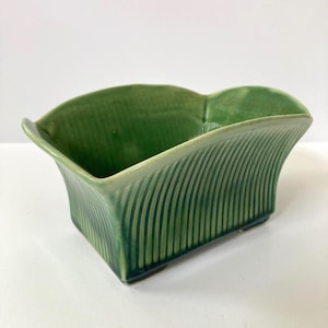 May include: A green ceramic planter with a rectangular shape and a curved, scalloped rim. The planter has vertical, ridged detailing on the sides and sits on small feet. The color is a vibrant, solid green.