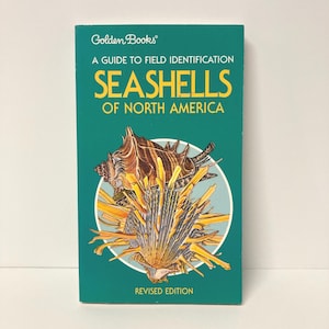 May include: A rectangular paperback book titled Golden Books A Guide to Field Identification Seashells of North America Revised Edition. The teal cover features a detailed illustration of two brown and yellow seashells centered in a white circle.