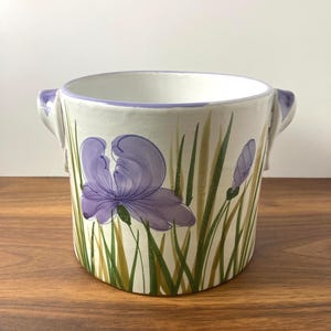 May include: A white ceramic planter with a hand-painted design of a purple iris flower and green grass. The planter has two small handles and a lavender rim. The design is on a cream-colored background.