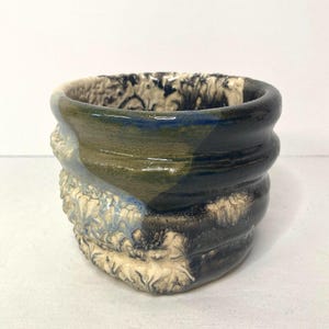 May include: A handmade ceramic cup with a textured exterior and a smooth, glazed interior. The cup features a color palette of blue, green, and black, with a unique, organic design. The rim is dark, and the base has a textured, almost floral pattern.
