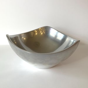 May include: A silver-colored, square-shaped bowl with gently curved sides. The bowl has a smooth, reflective surface and a modern design. This decorative bowl is suitable for serving food or as a decorative accent piece.