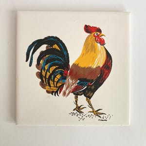 May include: A square ceramic tile featuring a colorful illustration of a rooster. The rooster has a red comb and wattle, yellow and brown body, and a black and blue tail. The rooster is standing on a white background.