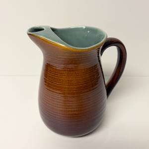 May include: A ceramic pitcher featuring a glossy, dark brown exterior with horizontal ribbed texture and a smooth, light blue interior glaze. The vessel includes a curved handle on the side and a wide, integrated pouring spout at the top rim.