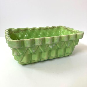 May include: A rectangular, light green ceramic planter with a textured, woven basket design. The rim has a scalloped edge with a brown trim. The planter is empty and sits on a white surface.