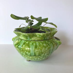 May include: A two-piece, green ceramic planter with a textured, speckled finish. The top section is a shallow dish, and the bottom is a rounded pot with three small feet. A small plant with green leaves is planted in the top section.