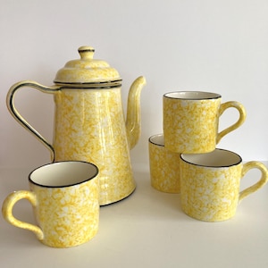 May include: A yellow enamelware coffee pot and four matching mugs. The set features a speckled yellow and white pattern with black trim. The coffee pot has a lid and a curved handle. The mugs have a rounded shape and a handle.