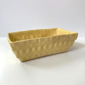 May include: A rectangular, pale yellow ceramic planter with a textured, diamond-patterned exterior. The planter has a slightly aged appearance, with subtle variations in color and texture, suggesting a vintage aesthetic. The interior is smooth and the edges are slightly rounded.