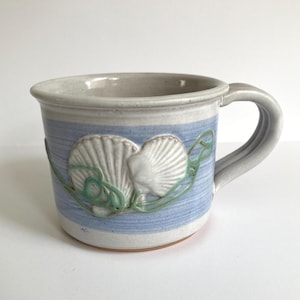 May include: Ceramic mug with a light blue and white striped design. The mug features a raised seashell design in white and green. The handle and rim are a light gray color. A decorative piece for the kitchen.