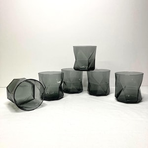 May include: Six gray, geometric-patterned drinking glasses. The glasses have a faceted design and a smoky gray color. One glass is lying on its side, while the others are upright. The glasses are arranged on a white surface.