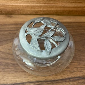 May include: A clear glass jar with a decorative metal lid. The lid features a bird and leaf design. The jar is round and sits on a wooden surface. The lid is silver in color.