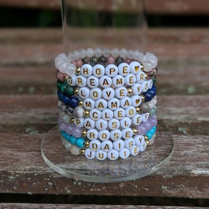 Könnte beinhalten: Eine Ansammlung von Armbändern mit Perlen, auf denen weiße Buchstaben Wörter und Namen bilden. Die Armbänder sind in verschiedenen Farben gehalten, darunter Rosa, Blau und Grün, und mit goldenen Perlen verziert. Die Armbänder werden auf einem transparenten, runden Ständer präsentiert.