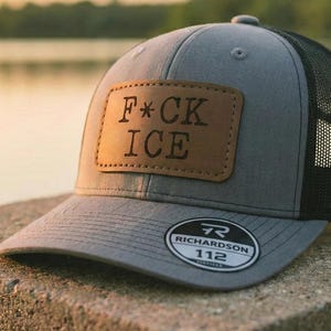 May include: Gray and black trucker hat with a leather patch that reads "F*CK ICE". The hat has a curved brim and a mesh back. The Richardson 112 logo is on the front.