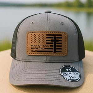 May include: Gray and black trucker hat with a leather patch featuring an American flag design and a cross. The patch includes the text "MAN OF GOD, HUSBAND, DAD". The hat has a curved brim and a Richardson 112 tag.