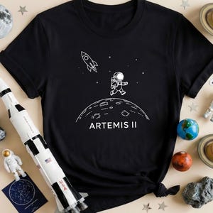 May include: Black t-shirt featuring a white graphic of an astronaut on a planet with a rocket and stars. The text "ARTEMIS II" is below the planet. The shirt is surrounded by space-themed toys, including a rocket, planets, and stars.