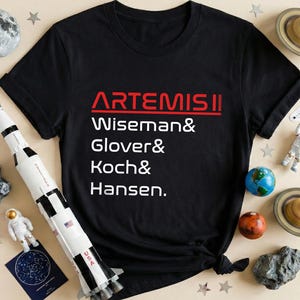 May include: Black t-shirt with the text "ARTEMIS II" in red and white, listing Wiseman, Glover, Koch, and Hansen. The image includes a toy rocket, planets, stars, and astronaut figurines, creating a space exploration theme.