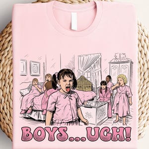 May include: Pink t-shirt featuring a cartoon illustration of girls in a bedroom, with the text "BOYS...UGH!". The graphic depicts several girls in pink dresses, with one girl sticking out her tongue. The design is printed on a light pink tee.