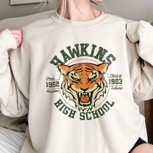 May include: A beige sweatshirt with a vintage-style graphic of a tiger and the text "Hawkins High School" with the years "1952" and "1983".