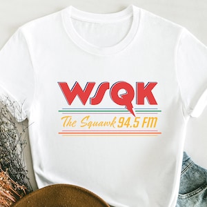 May include: White t-shirt with the text "WSQK The Squawk 94.5 FM" in red, green, orange, and yellow. The shirt has a rolled sleeve and is paired with denim shorts and a brown hat.