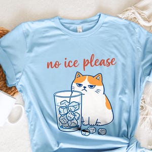 May include: Light blue t-shirt with the text "no ice please" in red. The shirt features a cartoon cat with an annoyed expression next to a glass of ice cubes. The cat is white and orange.