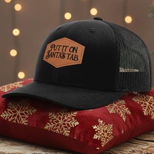 May include: A black trucker hat with a mesh back and a brown leather patch that reads "PUT IT ON SANTA'S TAB." The hat rests on a red velvet pillow with gold snowflake designs. The pillow is on a wooden stump.