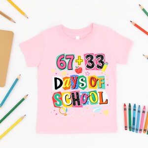May include: A light pink t-shirt with the text "67 + 33 Days of School" in a colorful, playful design. The graphic includes an apple, stars, and a pencil. Pencils and crayons are scattered around the shirt.