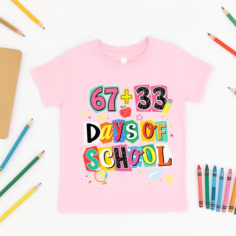 May include: A light pink t-shirt with the text "67 + 33 Days of School" in a colorful, playful design. The graphic includes an apple, stars, and a pencil. Pencils and crayons are scattered around the shirt.
