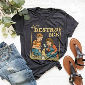 May include: Dark gray t-shirt featuring a retro illustration of two children with a magnifying glass and ice cube, with the text "Let's DESTROY ICE". The shirt is paired with blue jeans and brown sandals.