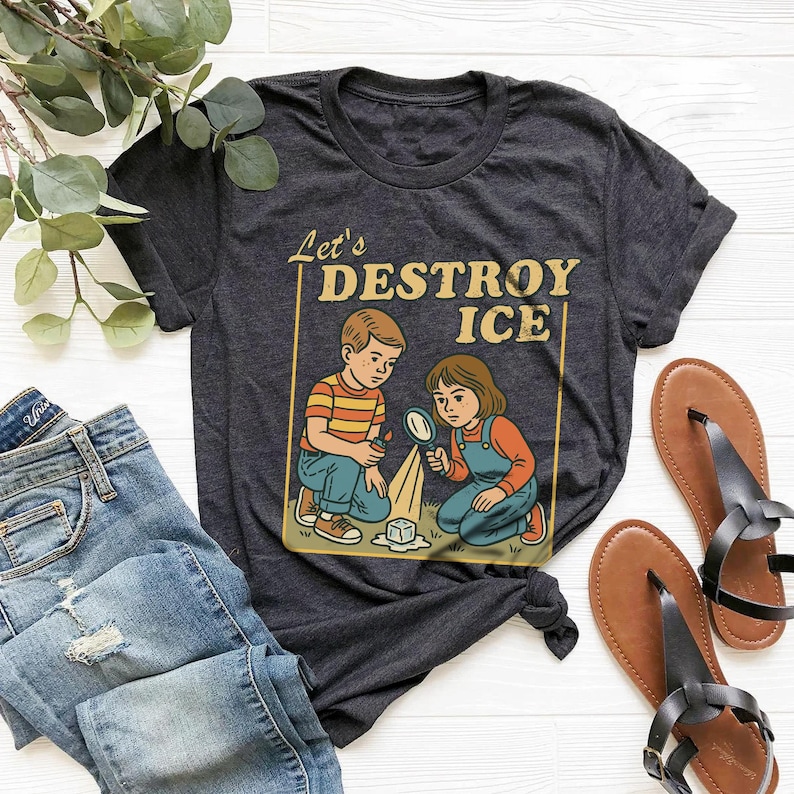 May include: Dark gray t-shirt featuring a retro illustration of two children with a magnifying glass and ice cube, with the text "Let's DESTROY ICE". The shirt is paired with blue jeans and brown sandals.