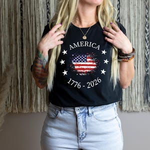 Może przedstawiać: Czarny t-shirt z postarzanym wzorem amerykańskiej flagi. Koszulka zawiera napis "AMERICA" nad flagą, otoczony gwiazdami, oraz daty "1776 - 2026" poniżej. Koszulka jest noszona z jasnoniebieskimi dżinsami.