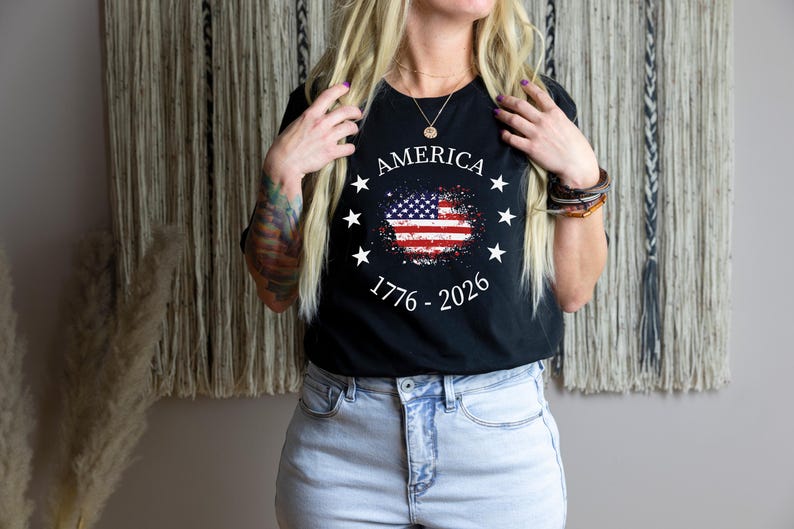 Może przedstawiać: Czarny t-shirt z postarzanym wzorem amerykańskiej flagi. Koszulka zawiera napis "AMERICA" nad flagą, otoczony gwiazdami, oraz daty "1776 - 2026" poniżej. Koszulka jest noszona z jasnoniebieskimi dżinsami.