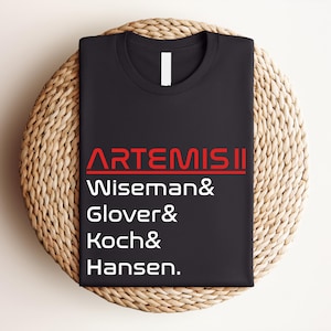 Pode incluir: Camiseta preta de gola redonda com o texto "ARTEMIS II" em vermelho e branco. Abaixo est&atilde;o os nomes Wiseman, Glover, Koch e Hansen. A camiseta est&aacute; dobrada e colocada sobre uma esteira redonda tecida.