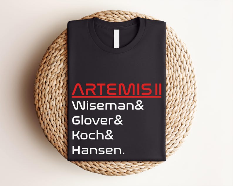 Pode incluir: Camiseta preta de gola redonda com o texto "ARTEMIS II" em vermelho e branco. Abaixo est&atilde;o os nomes Wiseman, Glover, Koch e Hansen. A camiseta est&aacute; dobrada e colocada sobre uma esteira redonda tecida.