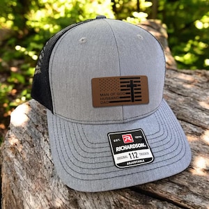May include: Gray trucker hat with a black mesh back and a brown leather patch. The patch features an American flag design with a cross and the words "MAN OF GOD, HUSBAND, DAD." The hat has a curved brim and an adjustable strap.