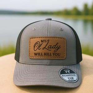 May include: Gray and black trucker hat with a leather patch. The patch has the text "MY Ol' Lady WILL KILL YOU" in a serif font. The hat has a curved brim and a Richardson 112 tag.