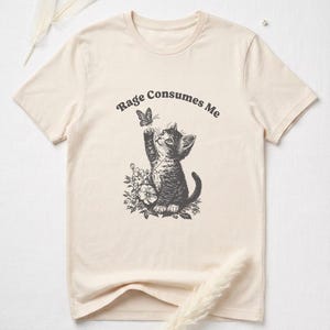 May include: Cream-colored t-shirt featuring a black and white illustration of a kitten reaching for a butterfly. The text "Rage Consumes Me" arches above the kitten. The kitten is surrounded by floral elements.