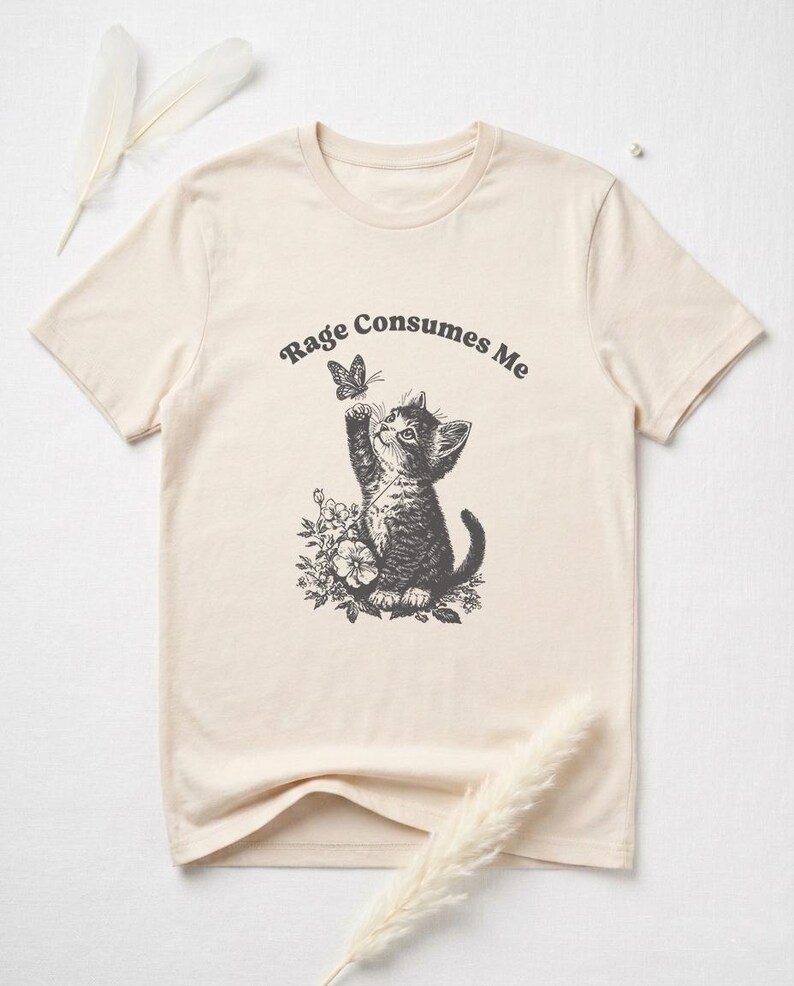 May include: Cream-colored t-shirt featuring a black and white illustration of a kitten reaching for a butterfly. The text "Rage Consumes Me" arches above the kitten. The kitten is surrounded by floral elements.