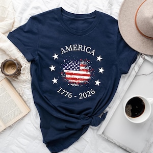 Może przedstawiać: Granatowa koszulka z nadrukiem amerykańskiej flagi w stylu vintage. Koszulka zawiera napis "AMERICA" w okręgu z gwiazdami, z latami "1776 - 2026" pod flagą. Na zdjęciu znajduje się r&oacute;wnież kapelusz, zegar i filiżanka kawy.