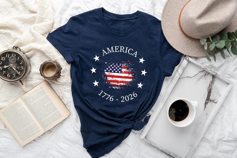 Może przedstawiać: Granatowa koszulka z nadrukiem amerykańskiej flagi w stylu vintage. Koszulka zawiera napis "AMERICA" w okręgu z gwiazdami, z latami "1776 - 2026" pod flagą. Na zdjęciu znajduje się r&oacute;wnież kapelusz, zegar i filiżanka kawy.