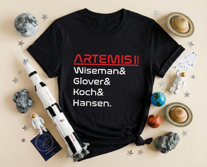 Pode incluir: Camiseta preta com o texto "ARTEMIS II" em vermelho e branco, listando Wiseman, Glover, Koch e Hansen. A imagem inclui um foguete de brinquedo, planetas, estrelas e figuras de astronautas, com tema de explora&ccedil;&atilde;o espacial.