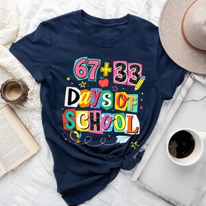 May include: Navy blue t-shirt with the text "67 + 33 Days of School" in colorful block letters. The design includes a pencil, apple, and stars. A vintage clock, open book, and coffee cup are also visible.