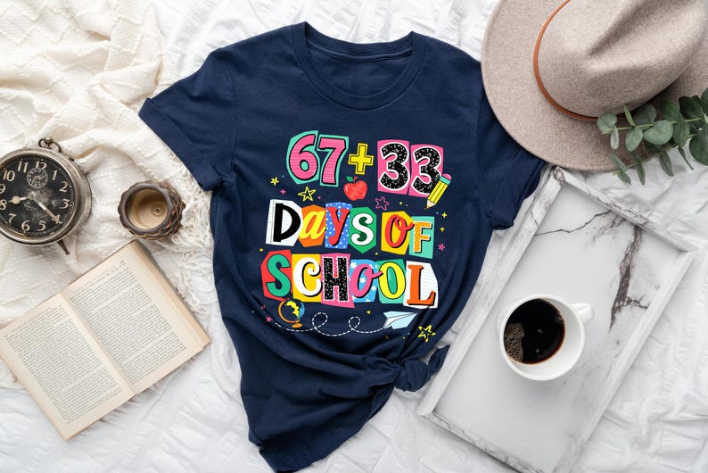 May include: Navy blue t-shirt with the text "67 + 33 Days of School" in colorful block letters. The design includes a pencil, apple, and stars. A vintage clock, open book, and coffee cup are also visible.