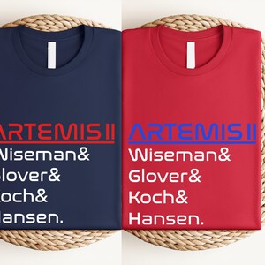 Pode incluir: Duas camisetas dobradas, uma azul marinho e outra vermelha, com o texto "ARTEMIS II" e os nomes Wiseman, Glover, Koch e Hansen impressos. As camisetas s&atilde;o exibidas em uma superf&iacute;cie circular tecida.