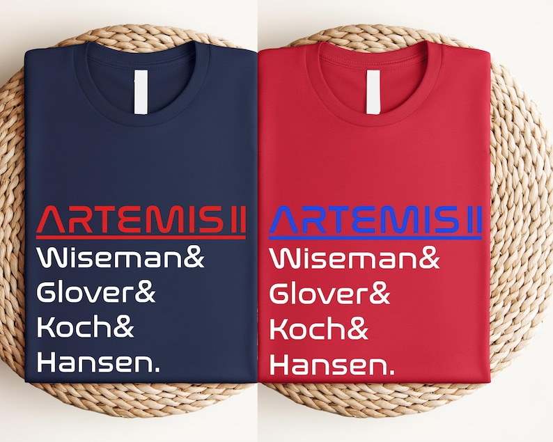Pode incluir: Duas camisetas dobradas, uma azul marinho e outra vermelha, com o texto "ARTEMIS II" e os nomes Wiseman, Glover, Koch e Hansen impressos. As camisetas s&atilde;o exibidas em uma superf&iacute;cie circular tecida.