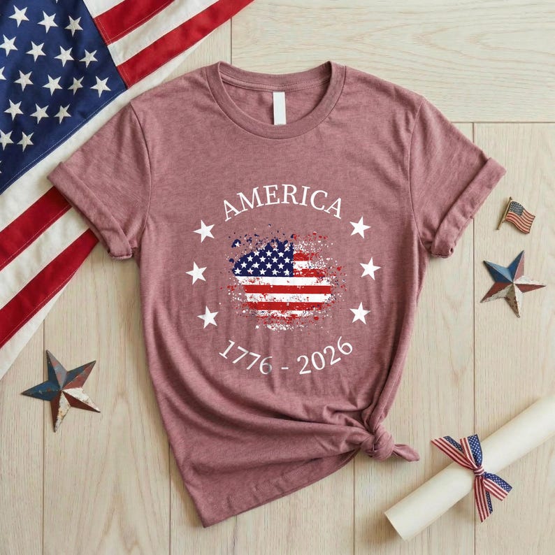 Może przedstawiać: Bordowa koszulka z napisem "AMERICA" nad zniszczonym wzorem amerykańskiej flagi. Koszulka zawiera r&oacute;wnież lata "1776 - 2026" i jest otoczona białymi gwiazdami. Amerykańska flaga i patriotyczne dekoracje znajdują się w tle.