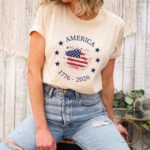Może przedstawiać: Kremowa koszulka z nadrukiem w postaci postarzonej flagi amerykańskiej. Nadruk zawiera napis "AMERICA" oraz lata "1776 - 2026". Projekt zawiera niebieskie gwiazdy i elementy flagi w kolorze czerwonym i niebieskim.