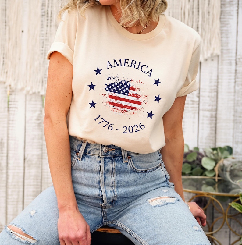 Może przedstawiać: Kremowa koszulka z nadrukiem w postaci postarzonej flagi amerykańskiej. Nadruk zawiera napis "AMERICA" oraz lata "1776 - 2026". Projekt zawiera niebieskie gwiazdy i elementy flagi w kolorze czerwonym i niebieskim.