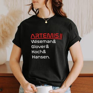 Pode incluir: T-shirt preto com a palavra "ARTEMIS II" em vermelho, acima dos nomes Wiseman, Glover, Koch e Hansen em branco. A t-shirt &eacute; de gola redonda.