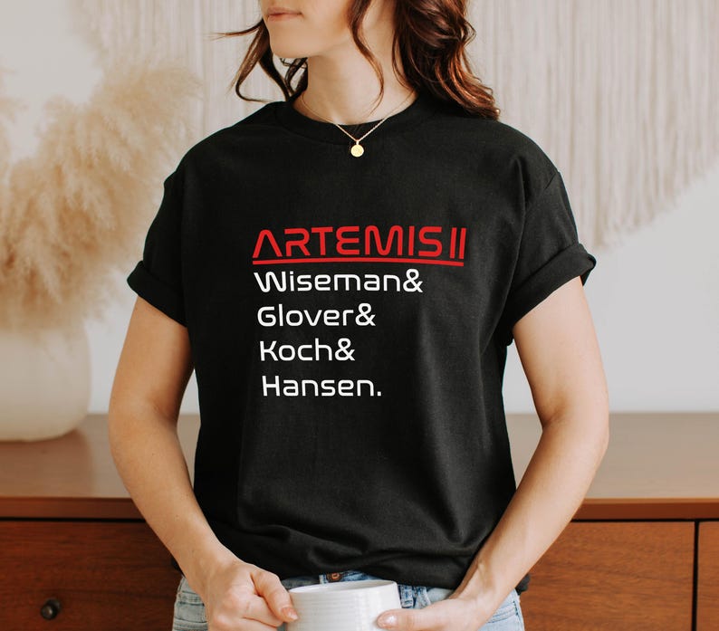 Pode incluir: T-shirt preto com a palavra "ARTEMIS II" em vermelho, acima dos nomes Wiseman, Glover, Koch e Hansen em branco. A t-shirt &eacute; de gola redonda.