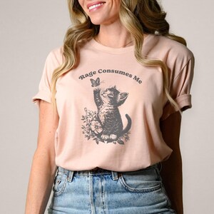 May include: A light pink t-shirt featuring a graphic of a kitten reaching for a butterfly, surrounded by floral elements. The text "Rage Consumes Me" is arched above the kitten. The shirt is made of soft material.